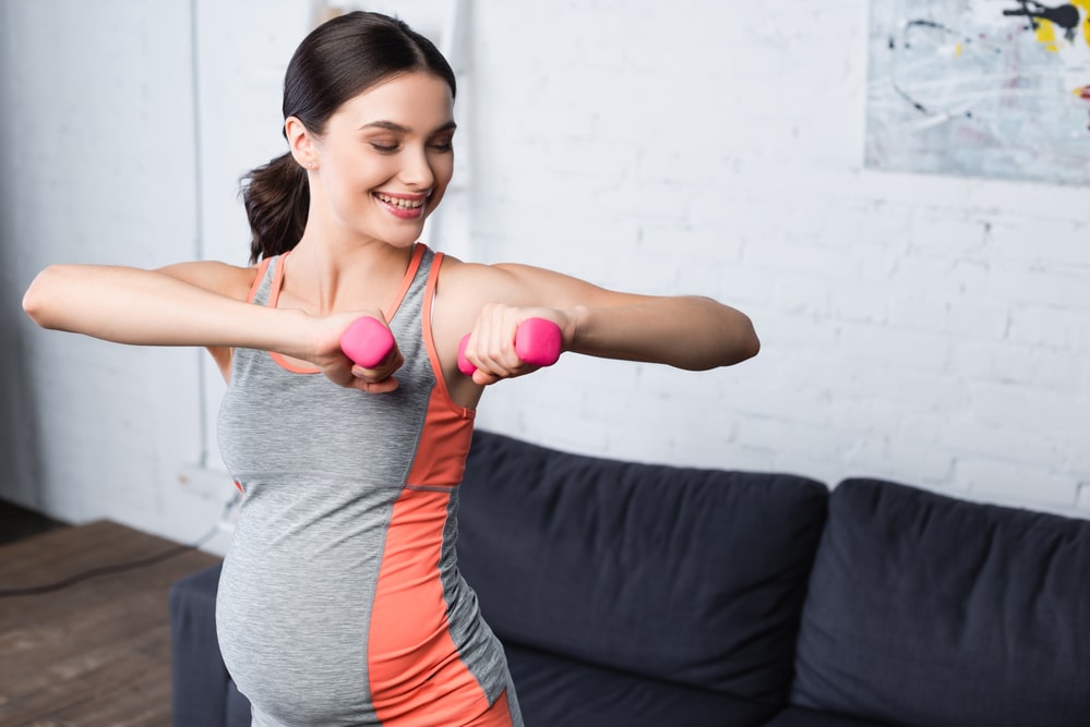 Pleased pregnant woman weightlifting pink dumbbells at home