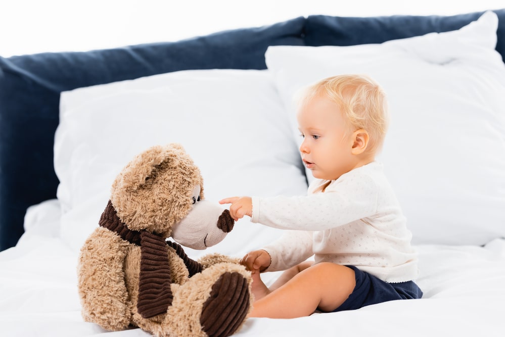 Toddler boy pointing at soft toy on bed on white background