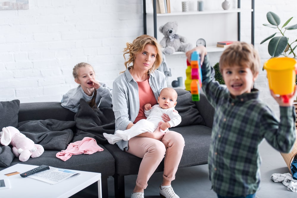 Tired mother with infant child sitting on couch while naughty siblings playing at home